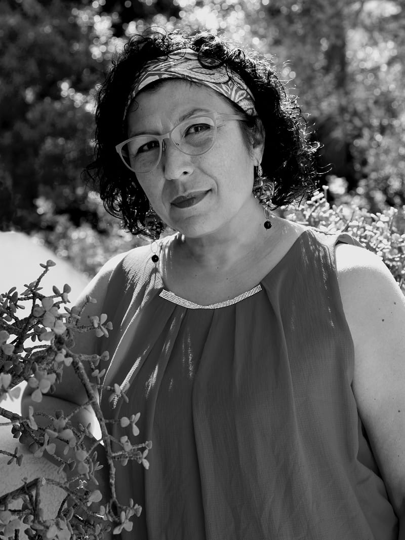 A black and white portrait of a woman outdoors, wearing a patterned headband, glasses, and a sleeveless top, with foliage in the background.