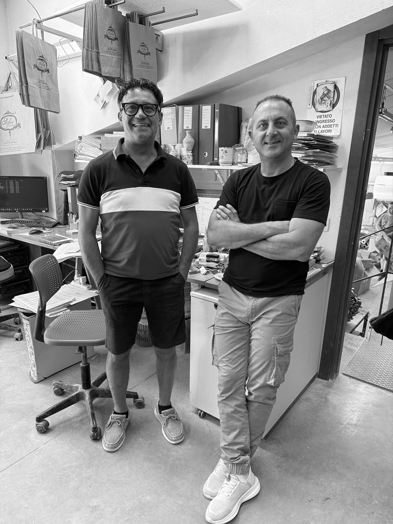 Two men standing in a workshop, wearing casual t-shirts and shorts. The photo is in black and white, showing one man in a dark striped polo shirt and shorts, and the other in a dark t-shirt and light cargo pants.