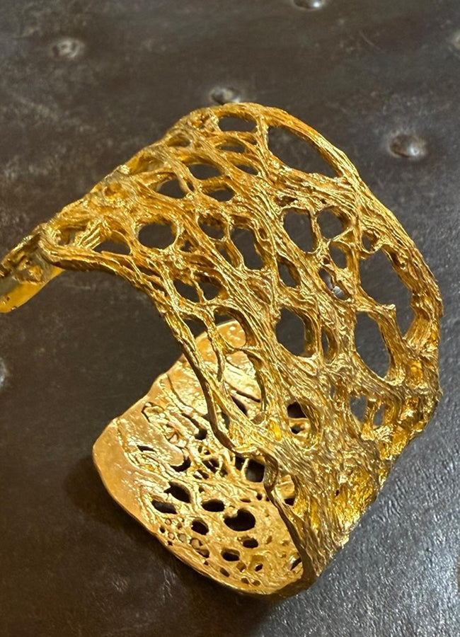 A textured gold cuff bracelet with an intricate openwork, tree-bark-like pattern, displayed on a dark, textured background.