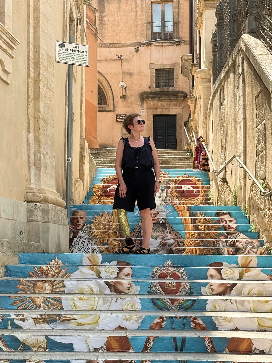 A woman in a black sleeveless top and shorts stands on a set of outdoor stairs painted with colorful classical-style illustrations, surrounded by historic beige stone buildings.