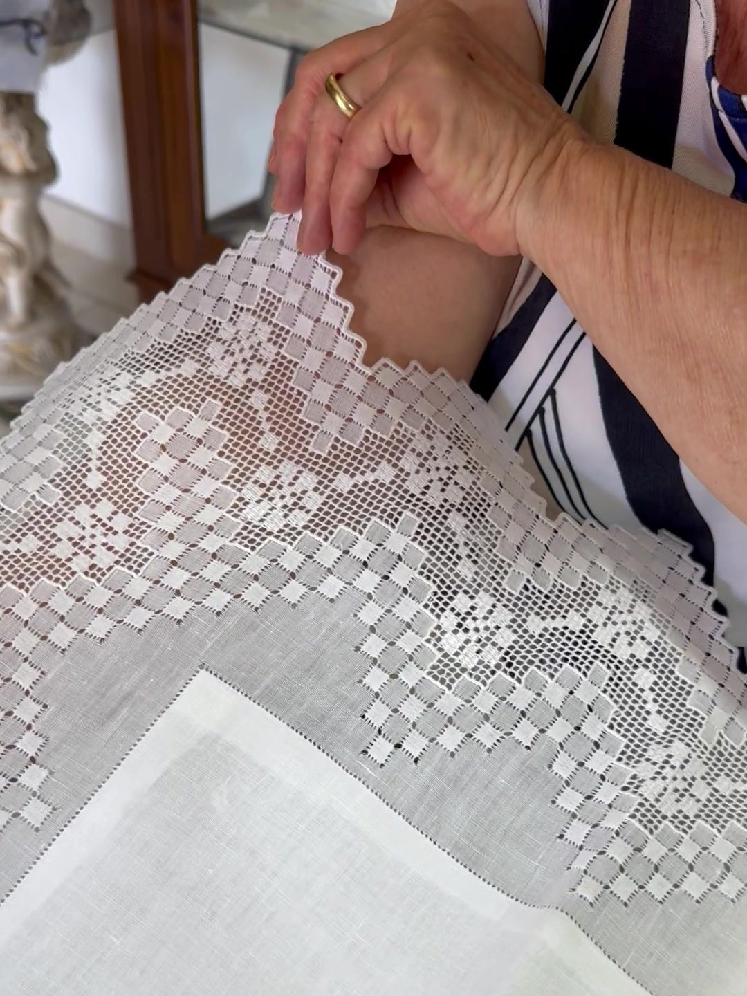 A close-up of a person's hands holding up a white embroidered lace textile, showing its detailed floral and geometric patterns. The person is wearing a black and white striped top and a gold ring, with a home interior background visible.