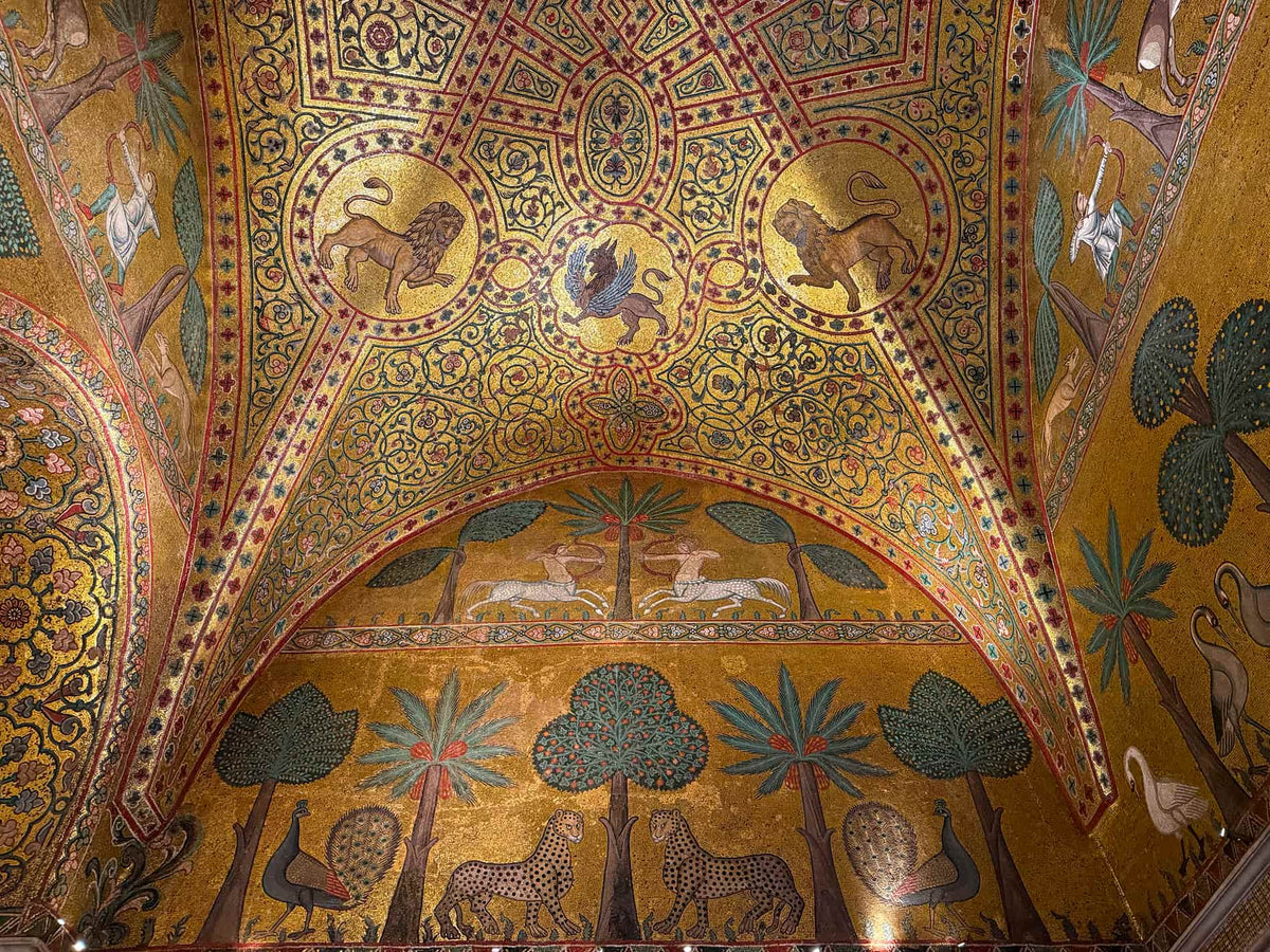 A close-up view of an ornate mosaic ceiling and wall featuring a golden background with intricate geometric patterns, lions, trees, birds, and animals in green, blue, red, and white tones.
