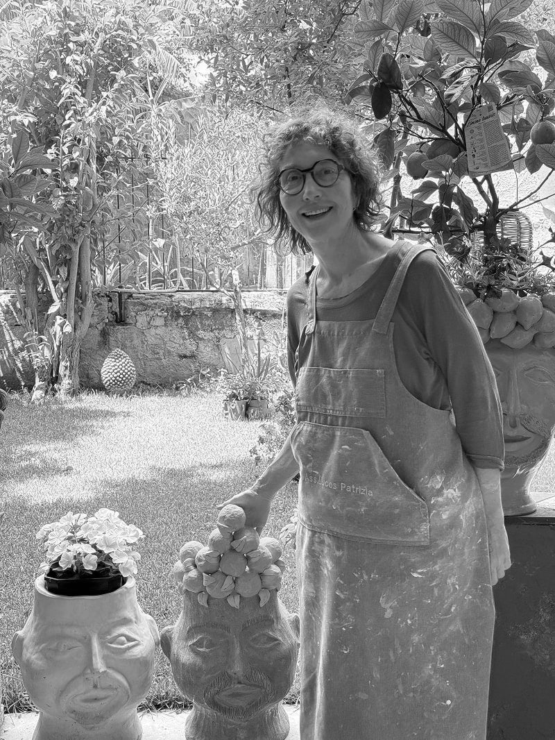 A woman wearing glasses and a clay-splattered apron stands in a garden next to two sculptural head planters, one filled with flowers.