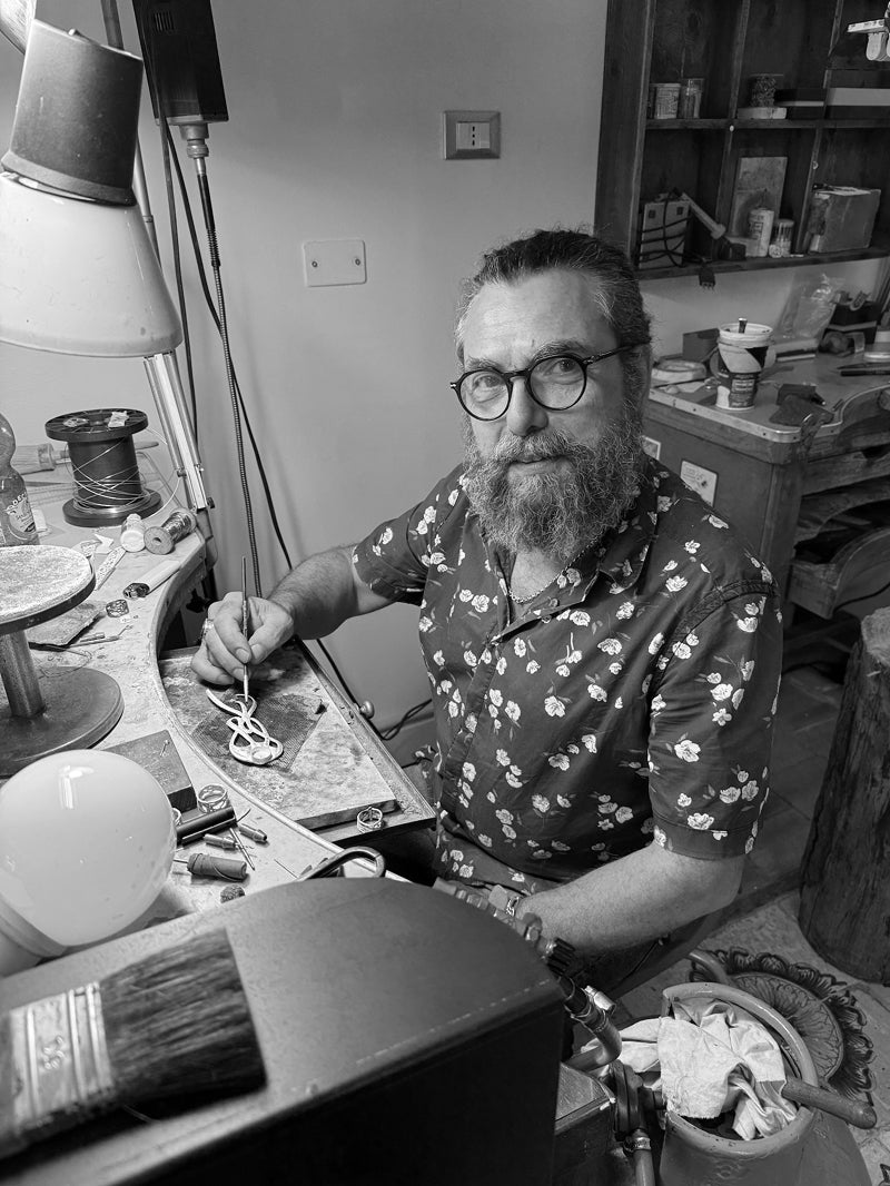 A black and white photograph of a bearded man with glasses wearing a short-sleeve button-up shirt with a small floral pattern, sitting at a metalsmith and jewelers workshop.