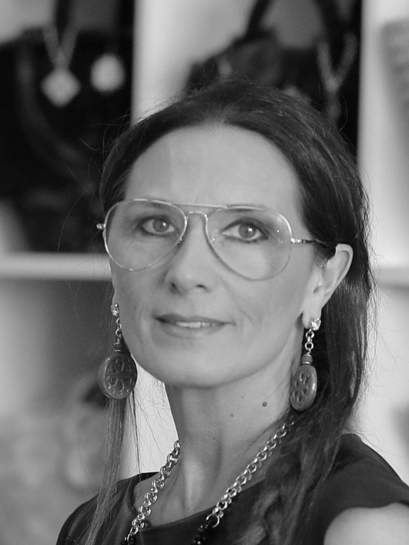 A black and white portrait of a woman with long dark hair, wearing aviator-style glasses, dangling earrings, and a chain necklace.