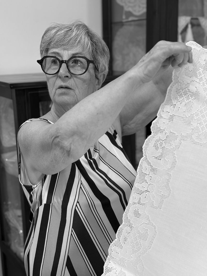 An older woman with short gray hair and thick-rimmed glasses holds up a white lace textile in a black-and-white indoor setting.