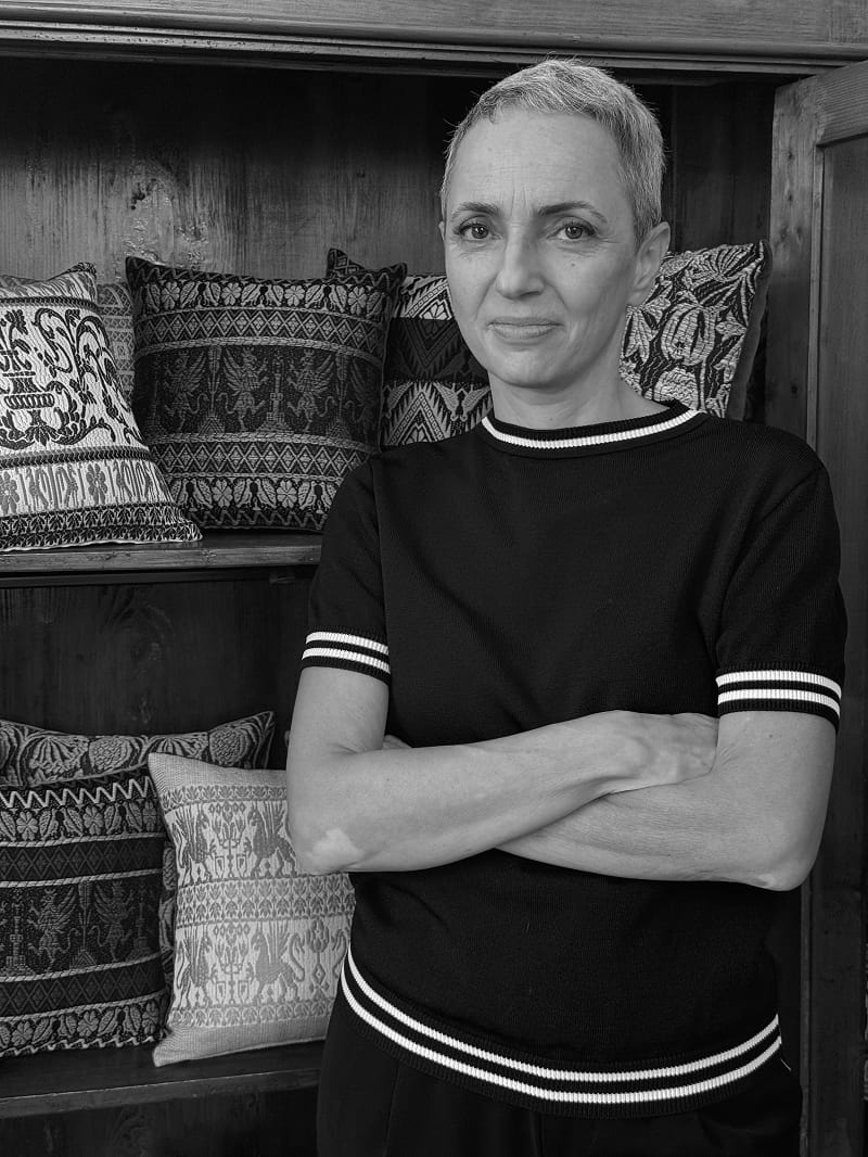 A black and white portrait of a woman with short hair, wearing a black short-sleeve top with white stripes on the collar, sleeves, and hem, standing in front of a wooden shelf with patterned pillows.