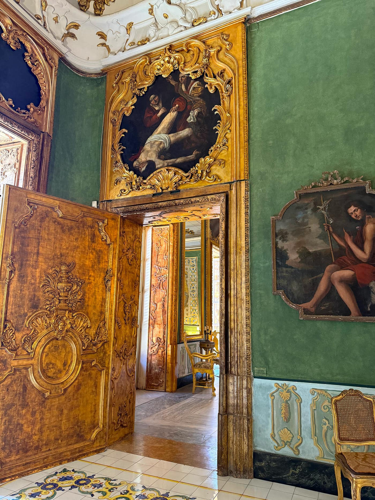 An ornate interior room with green walls, carved golden wood doors, framed classical paintings, and decorative molding. A large open wooden door dominates the left side, revealing another interior space beyond.