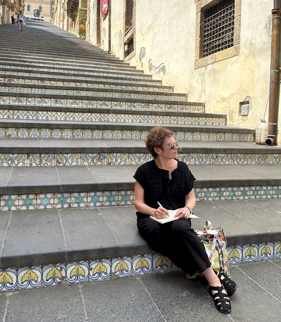 A woman wearing a black short-sleeve top and black pants sits on stone stairs with decorative blue, yellow, and white tile borders, holding a notebook and pen. She is in an outdoor urban setting with beige buildings and a narrow street in the background.