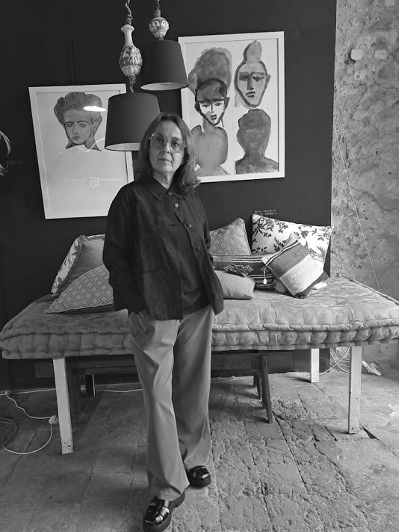 A black and white lifestyle photograph of a woman standing in a rustic room with a daybed, patterned pillows, and framed portrait paintings on the wall.