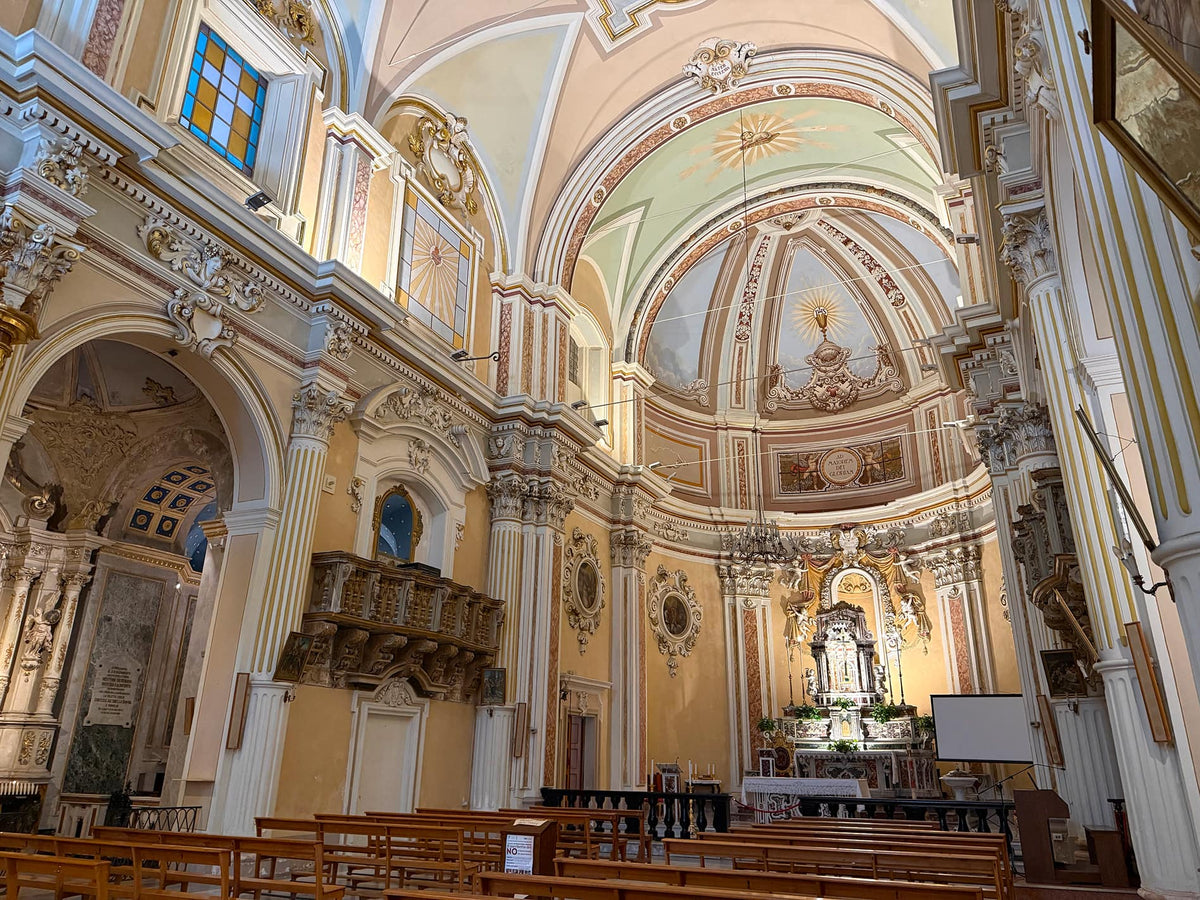 The interior of a grand, ornate church or cathedral with high arched ceilings, decorative frescoes, columns, pews, and an altar in the distance.