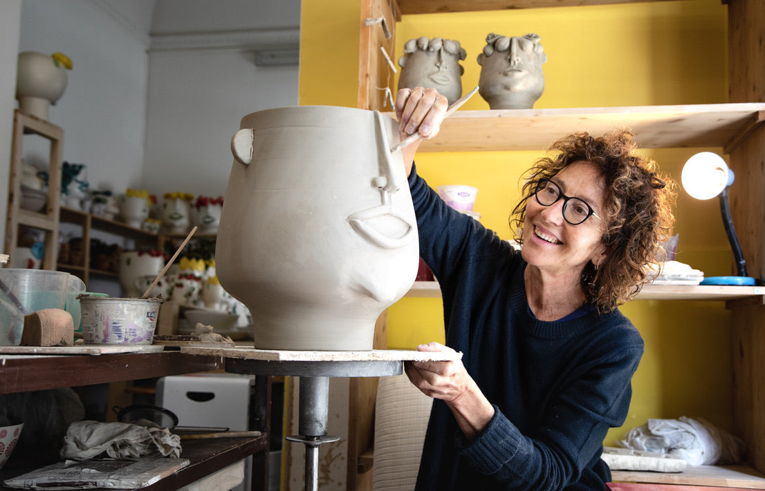 A woman with curly hair and glasses smiles while painting a large beige ceramic pot on a pottery wheel in a studio with shelves of pottery and a yellow wall.